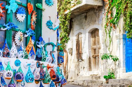 Bunte Keramikfiguren in verschiedenen Formen hängen an einer weißen Wand neben einem engen, sonnigen Gassenabschnitt mit blauen Türen und grünen Pflanzen.