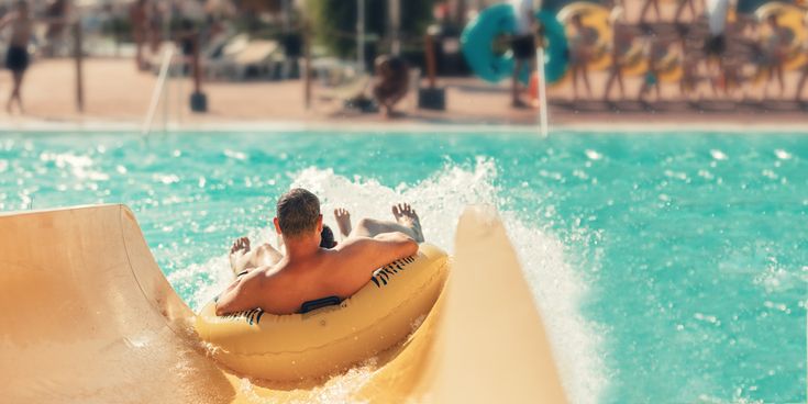 Erwachsener fährt mit Kind auf einem gelben Reifen eine Wasserrutsche hinunter und spritzt ins Becken