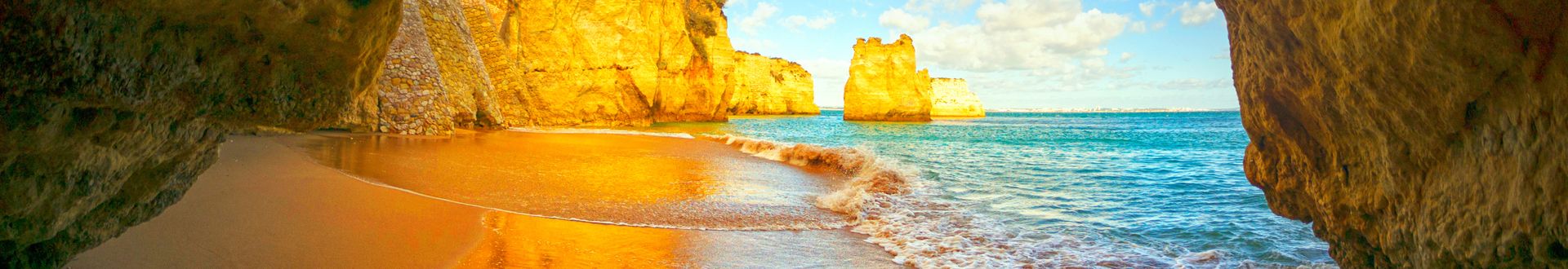 Blick auf einen Strand mit goldenem Sand und sanften Wellen, umgeben von hohen, gelben Felsen und blauem Himmel mit Wolken.
