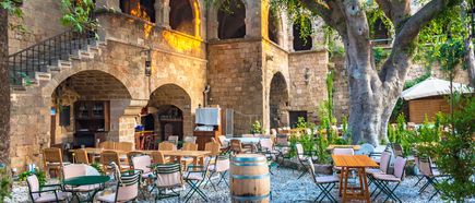 Innenhof einer Taverne mit steinernen Bögen, mehreren Tischen und Stühlen sowie einem großen Baum in der Mitte.