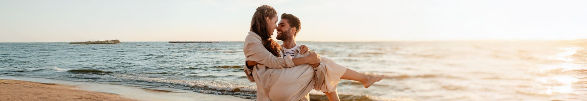 Mann trägt seine Partnerin am Strand im Sonnenuntergang, beide lächeln sich verliebt an