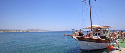 Mehrere Personen sitzen auf einem hölzernen Segelboot, das an einem Steg im Meer vor einer Küstenlandschaft festgemacht ist.
