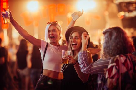 Drei junge Frauen feiern ausgelassen auf einer Party, halten Getränke in der Hand und lachen inmitten einer Menschenmenge mit bunter Beleuchtung