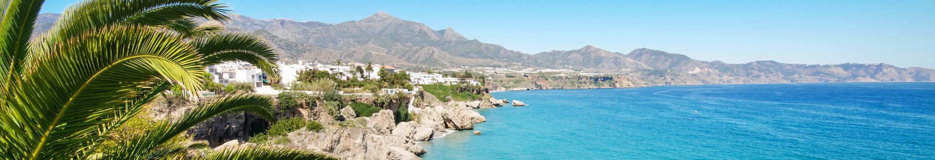 Küstenblick auf Nerja mit Palmen im Vordergrund, weißen Häusern auf Felsen und den Bergen Andalusiens im Hintergrund