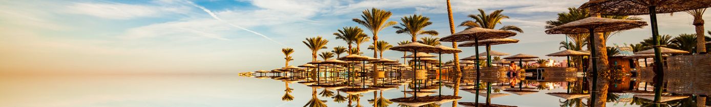 Spiegelglatte Wasseroberfläche reflektiert Palmen und Strohdach-Sonnenschirme unter blauem Himmel mit leichten Schleierwolken in Hurghada, Ägypten.