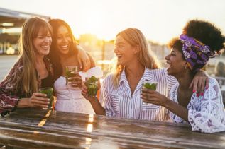 Vier junge Frauen sitzen lachend an einem Holztisch im Sonnenuntergang und halten Cocktails in der Hand