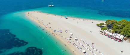 Luftaufnahme eines sandigen Strandes mit Liegestuhlreihen, Sonnenschirmen, Badegästen und mehreren Booten im türkisfarbenen Meer