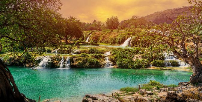 Mehrstufiger Wasserfall fließt in türkisfarbenen See, umgeben von grünen Bäumen und Hügeln bei Sonnenuntergang