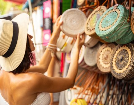 Frau mit Sonnenhut und weißem Oberteil betrachtet runde geflochtene Taschen an einem Marktstand
