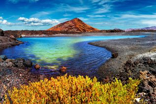 Vulkanische Landschaft mit einem kegelförmigen Berg, einem türkisfarbenen See und gelb-grünem Busch im Vordergrund