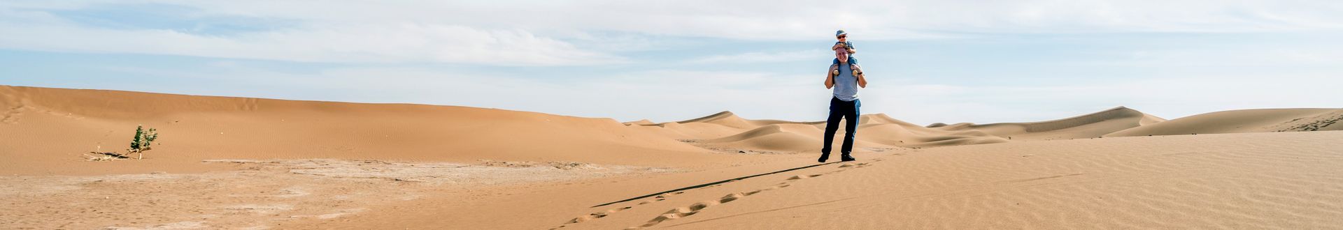 Ein Mann trägt ein Kind auf den Schultern und läuft durch eine weite Wüstenlandschaft mit Dünen unter blauem Himmel