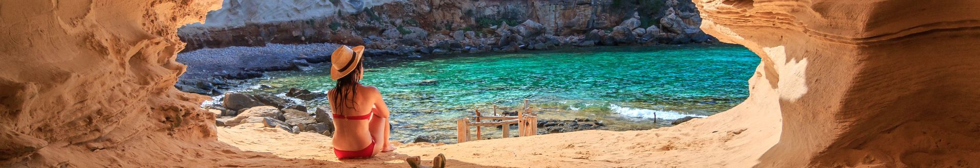 Frau in rotem Bikini und Strohhut sitzt in einer Felsenhöhle am Strand von Cala en Baster auf Formentera und blickt auf das türkisfarbene Meer