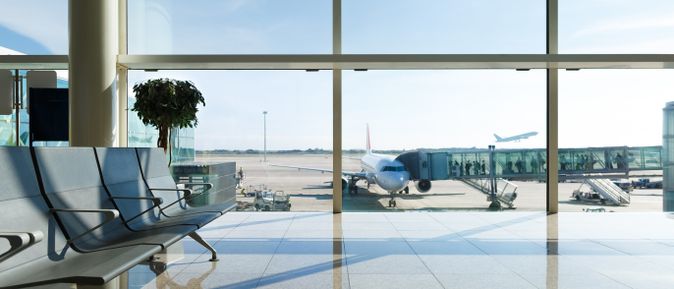 Moderne Wartezone im Flughafenterminal mit Blick auf das Vorfeld, ein angedocktes Passagierflugzeug an der Fluggastbrücke sowie ein startendes Flugzeug im Hintergrund