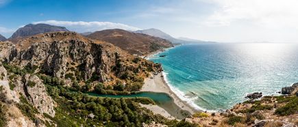 Küstenlandschaft mit felsigen Hügeln, grünem Flusslauf und sandigem Strand an türkisblauem Meer unter bewölktem Himmel.