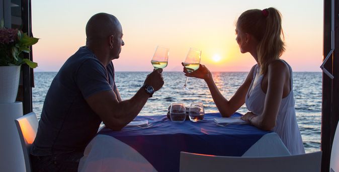 Ein Paar sitzt in einem Restaurant direkt am Meer bei Sonnenuntergang und halten Weingläser