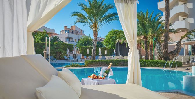 Poolbereich des Hotels Girasol auf Mallorca mit einem Balibett, Tisch, Sektflasche, Gläsern und Palmen im Hintergrund.