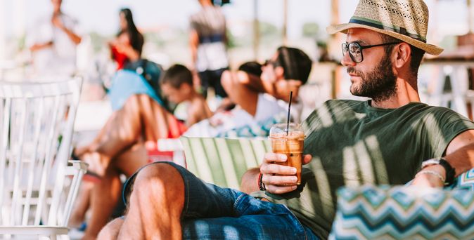 Mann mit Strohhut und grünem T-Shirt sitzt entspannt auf einer Terrasse und hält ein Getränk mit Strohhalm in der Hand.
