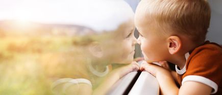 Kind lehnt am Fenster eines fahrenden Fahrzeugs und blickt auf die vorbeiziehende Landschaft im warmen Sonnenlicht