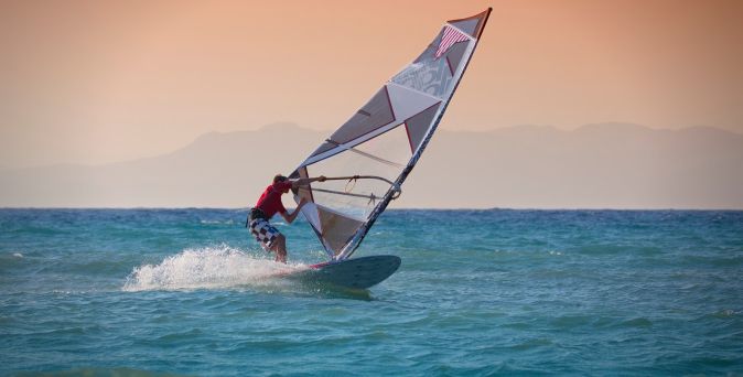 Person beim Windsurfen auf dem Meer mit buntem Segelbrett und Spritzwasser vor orangefarbenem Himmel