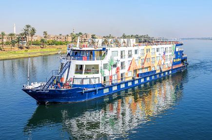 Das Nilkreuzfahrtschiff MS Nile Excellence mit weißem und blauem Rumpf, mehreren Decks und dekorativen Mustern fährt auf dem Nil mit Ufer und Palmen im Hintergrund