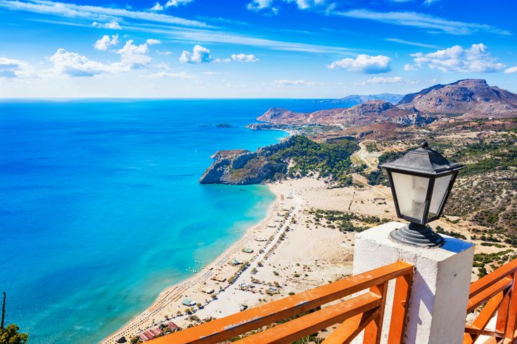 Blick von einer Terrasse mit Holzgeländer und Laterne auf den Tsambika-Strand mit türkisblauem Meer und bewölktem Himmel auf Rhodos.