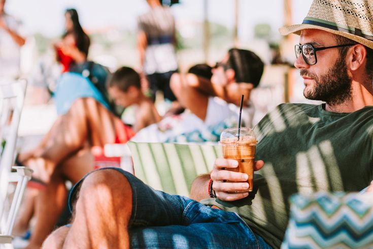 Mann mit Strohhut und grünem T-Shirt sitzt entspannt auf einer Terrasse und hält ein Getränk mit Strohhalm in der Hand.
