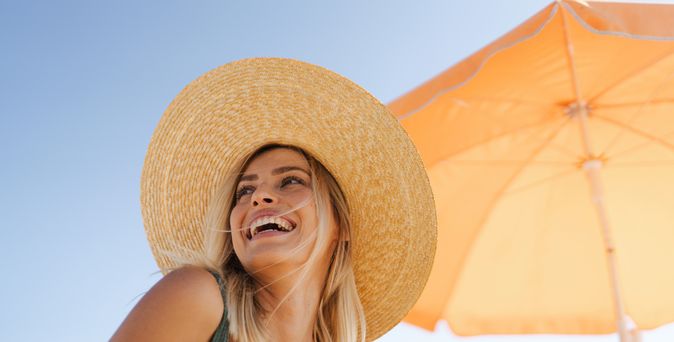 Frau mit großem, geflochtenem Strohhut sitzt unter einem orangefarbenen Sonnenschirm an einem sonnigen Tag