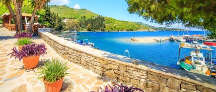 Blick auf den Hafen von Kouloura auf Korfu mit einer Steinmauer, bepflanzten Töpfen, mehreren Booten im Wasser und grünen Hügeln im Hintergrund