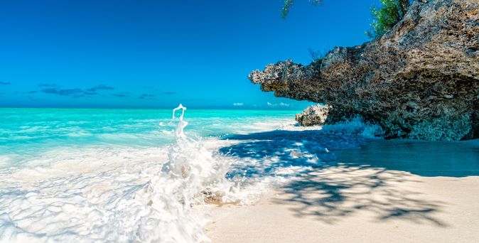 Wellen schlagen an einen weißen Sandstrand unter einem Felsen mit klarem blauem Himmel und türkisfarbenem Meer