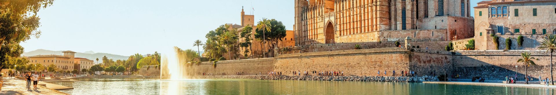 Panoramablick auf die Kathedrale La Seu in Palma de Mallorca mit Springbrunnen, Wasserbecken und Spaziergängern entlang der Uferpromenade