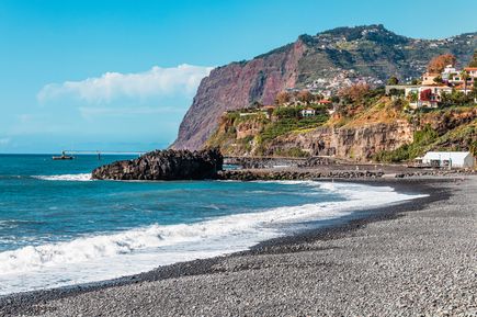 Kiesstrand Praia Formosa auf Madeira mit Atlantikwellen, dunklen Felsen, Küstenstraße und steilen Berghängen mit Häusern im Hintergrund.