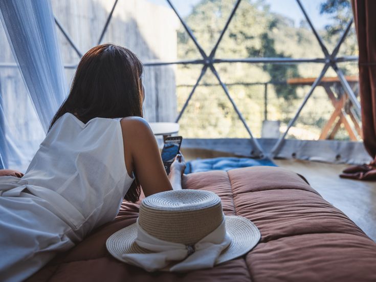 Person liegt auf einem Bett in einem Glamping-Dome und blickt auf ein Smartphone; im Vordergrund liegt ein Strohhut, im Hintergrund sind große Panoramafenster mit Blick ins Grüne zu sehen.