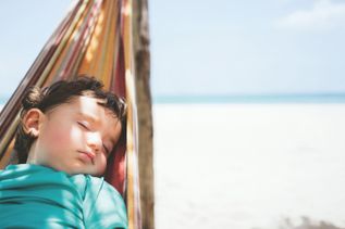 Junge liegt entspannt in einer bunten Hängematte am Strand, mit Blick auf den hellen Sand und das ruhige, türkisfarbene Meer unter klarem Himmel