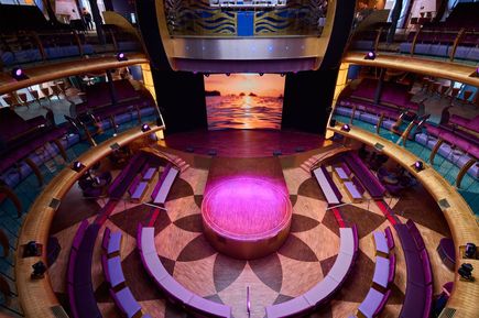 Rundbühne mit pink beleuchtetem Podest in einem mehrstöckigen Theatersaal mit halbkreisförmigen Sitzreihen und einem großen Bildschirm im Hintergrund, der einen Sonnenuntergang über dem Meer zeigt