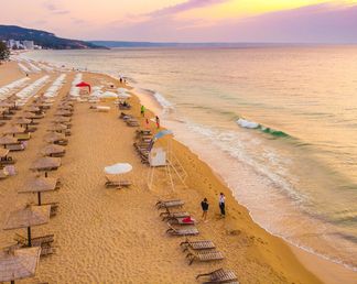 Strand von Goldstrand, Bulgarien, mit Liegen und Sonnenschirmen, zwei Personen am Wasser, sanfte Wellen, Sonnenuntergang im Hintergrund.