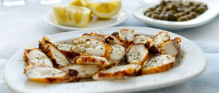 Gegrillte Tintenfischstücke auf einem weißen Teller mit Zitronenspalten und einer Schale Kapern im Hintergrund