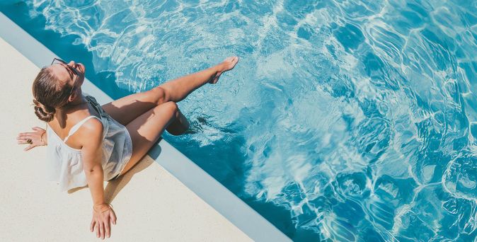 Person sitzt am Rand eines Swimmingpools und lässt die Beine im klaren blauen Wasser baumeln, während Sonnenreflexionen und Wellenmuster auf der Wasseroberfläche sichtbar sind.