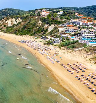 Luftaufnahme eines langen Sandstrands mit Reihen von Sonnenschirmen und Liegen, dahinter bewaldete Hügel und Gebäude.
