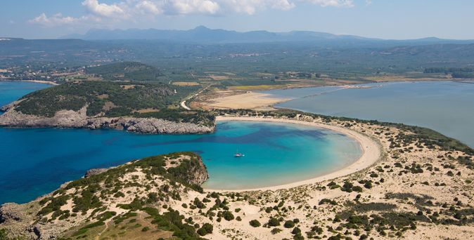 Luftaufnahme einer geschwungenen Sandbucht mit klarem blauem Wasser und bewachsenen Dünen im Vordergrund.