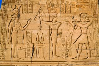 Hieroglyphen und Reliefs an der Wand des Kom-Ombo-Tempels zeigen drei ägyptische Götterfiguren mit traditionellen Kopfbedeckungen und Symbolen