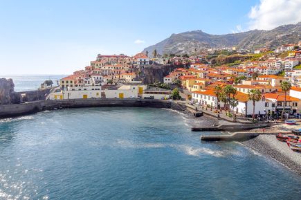 Blick auf die Küstenstadt Funchal auf Madeira mit orangefarbenen Dächern, Uferpromenade, kleiner Hafenbucht, Fischerbooten und terrassenförmig angelegten Berghängen im Hintergrund