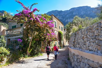 Malerische Gasse im Dorf Deià auf Mallorca mit Natursteinhäusern, blühender Bougainvillea und Wanderern vor Bergkulisse
