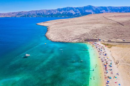 Luftaufnahme der Plaža Ručica auf der Insel Pag in Kroatien mit türkisfarbenem Meer, hellem Kiesstrand, Badenden und der charakteristischen kargen Küstenlandschaft der Adria