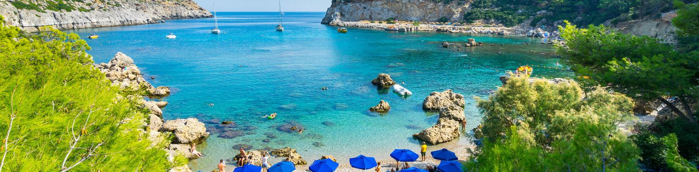 Bucht mit klarem Wasser, Felsen und bewachsenem Hügel im Hintergrund, Strand mit blauen Sonnenschirmen und Menschen.