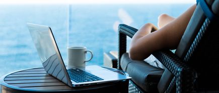 Frau sitzt entspannt auf dem Balkon eines Kreuzfahrtschiffs in einem Lounge‑Sessel, mit einem geöffneten Laptop und einer Tasse auf dem kleinen Tisch, während im Hintergrund das Meer zu sehen ist