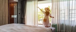 Frau in weißem Kleid und Strohhut steht vor bodentiefen Fenstern in Hotelzimmer und blickt auf Palmen draußen