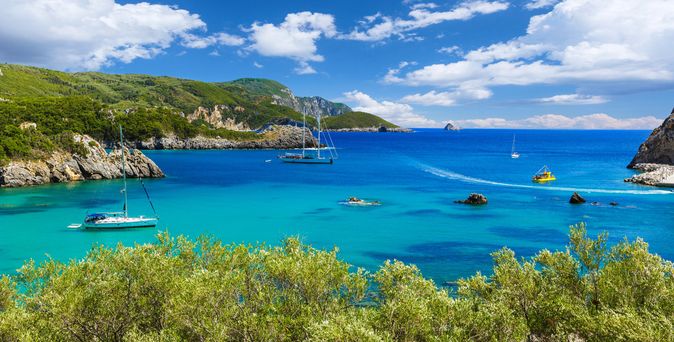 Bucht mit türkisfarbenem Wasser, Segel- und Motorboote, grüne Hügel und bewölkter Himmel auf Korfu