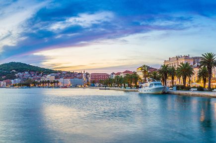 Blick auf die Uferpromenade und den Hafen von Split in Kroatien mit Palmen, historischen Gebäuden, einer Yacht am Kai und ruhigem Wasser in der Abenddämmerung