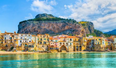 Blick auf die Küstenstadt Cefalù in Sizilien mit historischen Gebäuden am Strand, türkisfarbenem Meer und dem markanten Felsen La Rocca im Hintergrund