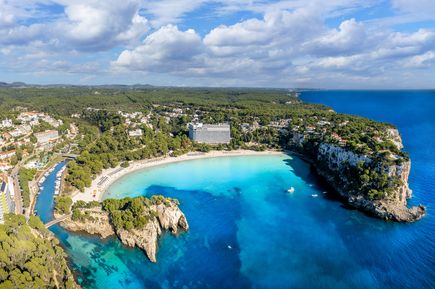 Die Bucht Cala Galdana auf Menorca zeigt türkisblaues Wasser, einen Sandstrand und Hotels, die von grünen Hügeln umgeben sind